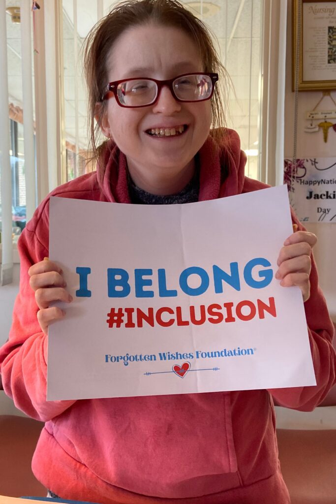 Nicole is a women who is sight impaired. She is holding a sign that says I Belong and smiling.